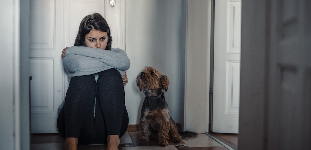 A woman Is Sitting Exhausted On The Floor of a House With Her Dog Next To Her. She looks sad and appears to be staring blanky ahead of her.