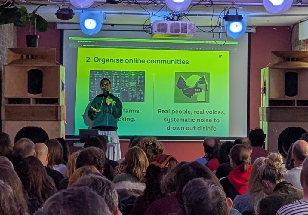 A presenter speaks on stage in front of a green slide labelled “2. Organise online communities.”
Two images appear on the slide: one of computer servers with the caption “Bots & troll farms, hashtag hacking,” and another of shaking hands with the text “Real people, real voices, systematic noise to drown out disinfo.”
The audience watches attentively in the event space.