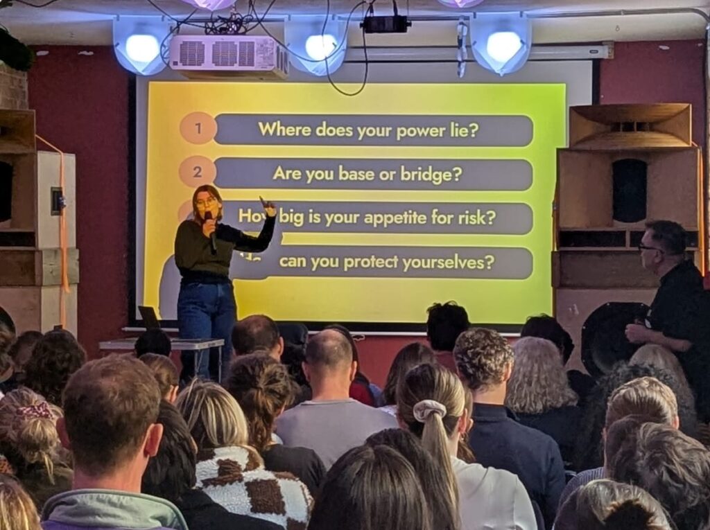 A speaker stands on stage holding a microphone and gesturing toward a large yellow slide that reads:
Where does your power lie?
Are you base or bridge?
How big is your appetite for risk?
How can you protect yourselves?
They face an attentive seated audience in a warmly lit room with brick walls and wooden speakers.