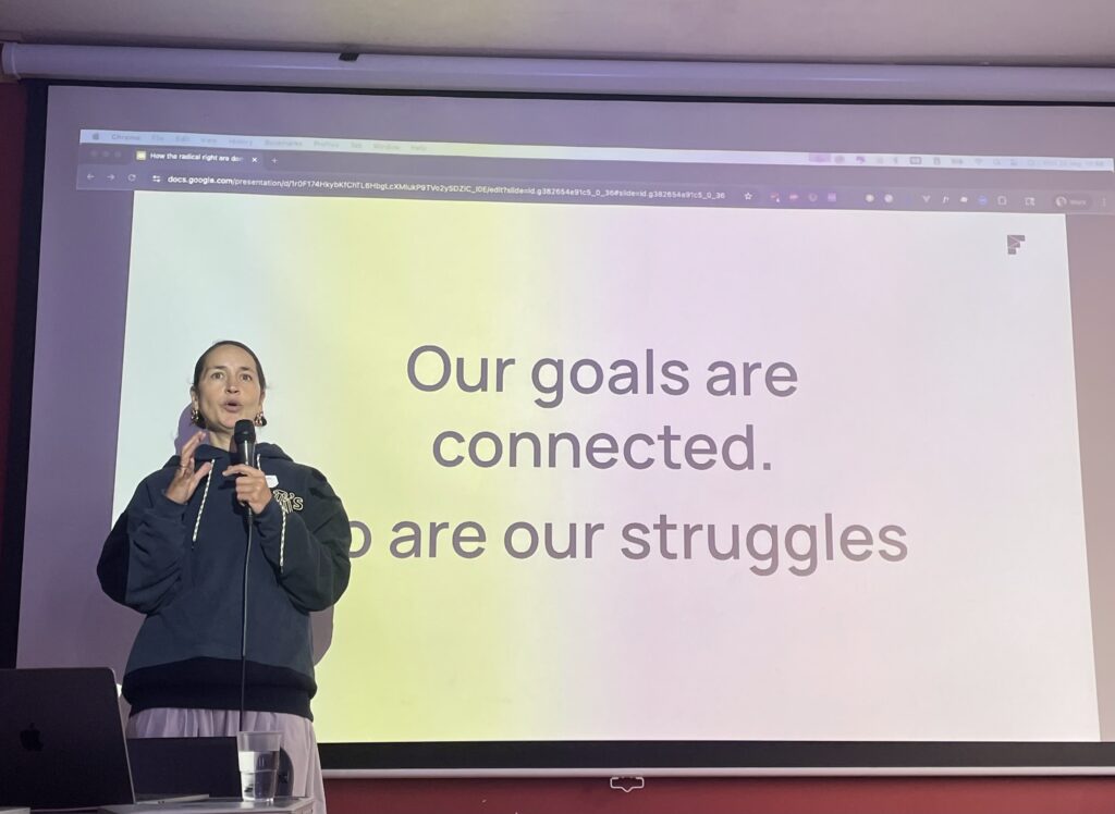 A speaker stands on stage holding a microphone, addressing an audience in front of a large slide that reads, “Our goals are connected. So are our struggles.”