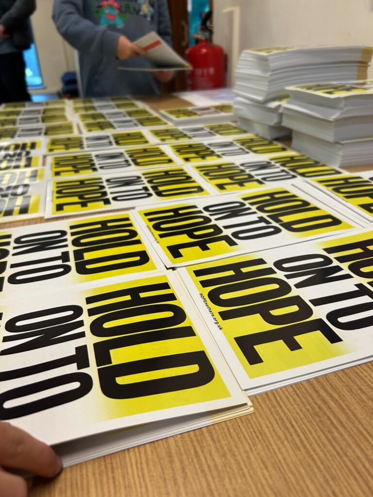 Stacks of yellow-and-black "Hold on to Hope" leaflets spread across a table, while volunteers assemble or sort them.