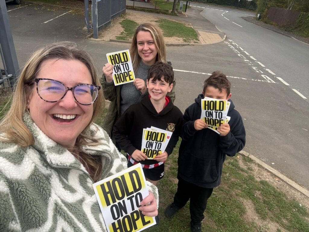 Two smiling women and two children stand outdoors holding "Hold on to Hope" leaflets, posing for a cheerful group photo.
