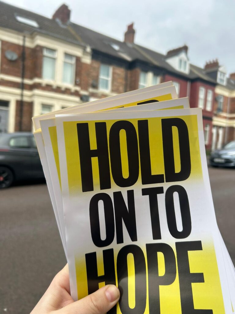 A hand holds several yellow-and-black leaflets reading "Hold on to Hope," with terraced houses blurred in the background.