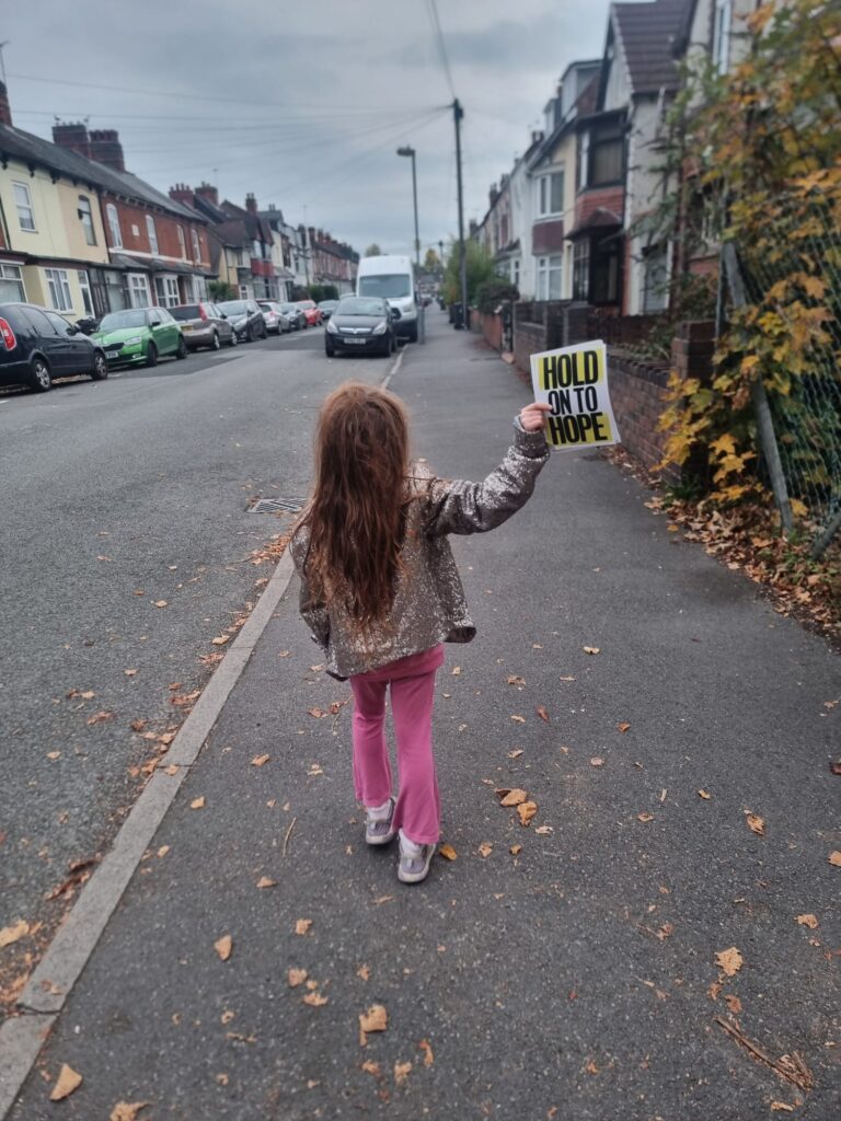 A young child with long hair and a sparkly jacket walks down a residential pavement holding a bright yellow-and-black poster that says "Hold on to Hope."