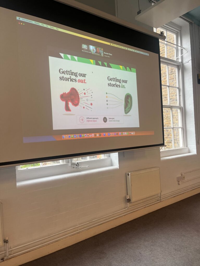 Projected presentation slide in a workshop space showing the phrases “Getting our stories out.” beside a megaphone graphic and “Getting our stories in.” beside an ear graphic, illustrating different approaches to climate campaigning and public engagement during a Zoom presentation displayed on a large screen.