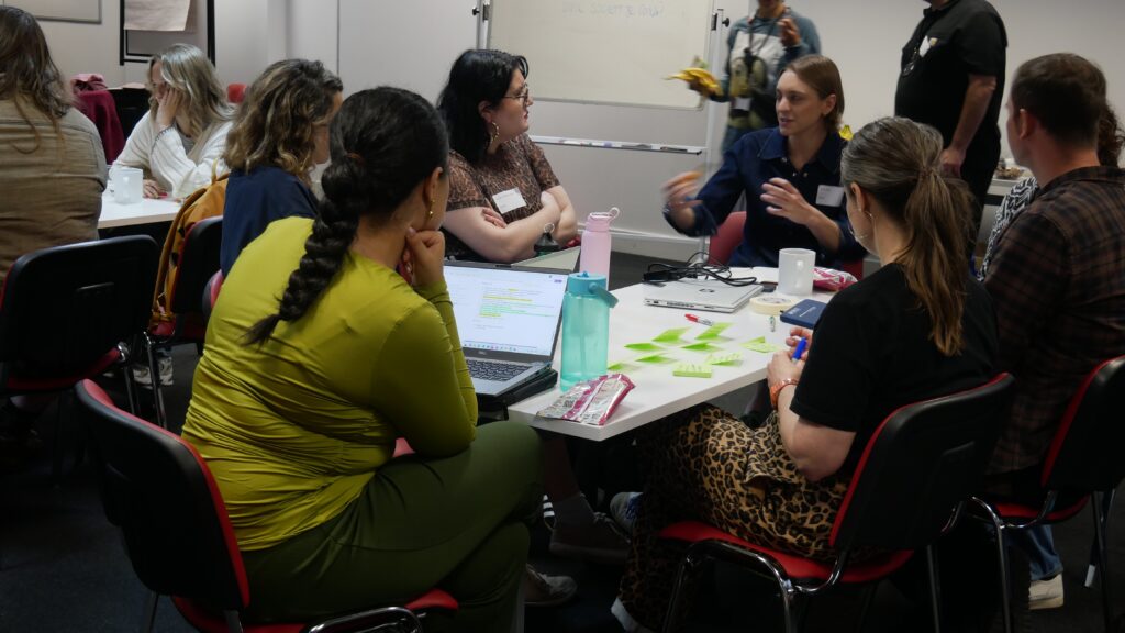 Charity and campaign staff workshop ideas around a table at the How We Win event. 6 participants are looking at one person who is speaking.