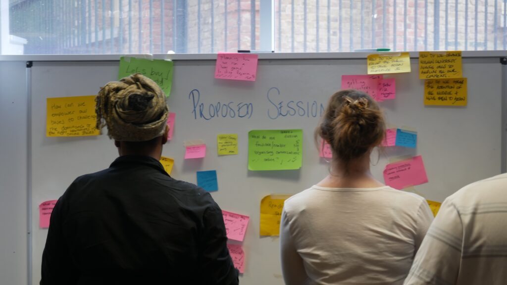 Two attendees at a Forward Action and Rally event have their backs to the camera as they review proposed session ideas on a whiteboard covered in sticky notes.