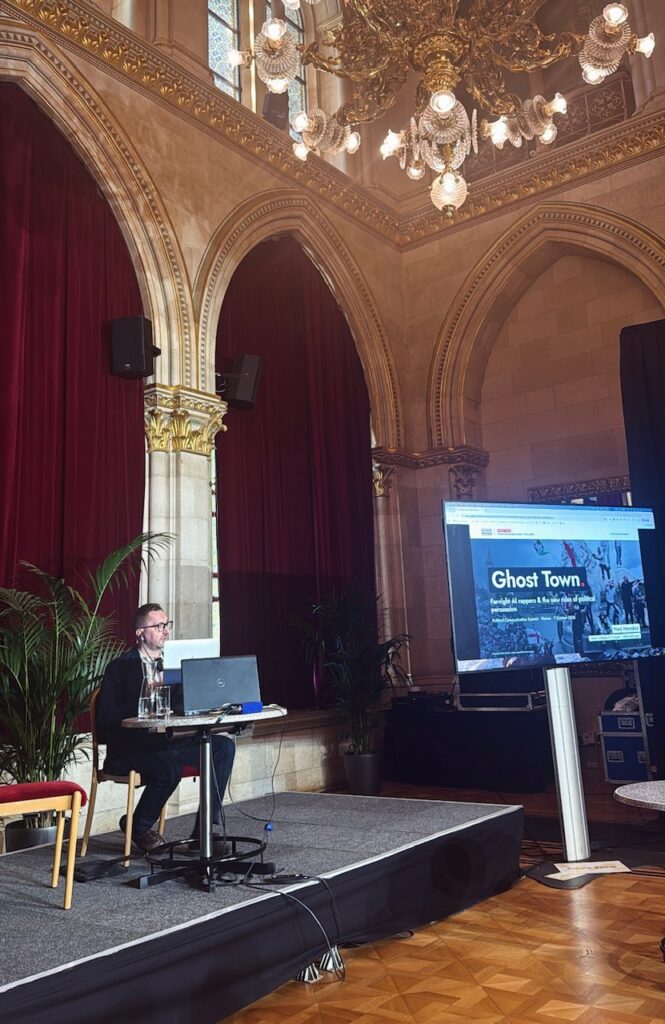Ned Mendez of Alliance4Europe presenting “Ghost Town: Far-right AI rappers and the new rules of political persuasion” in an ornate hall with red curtains.