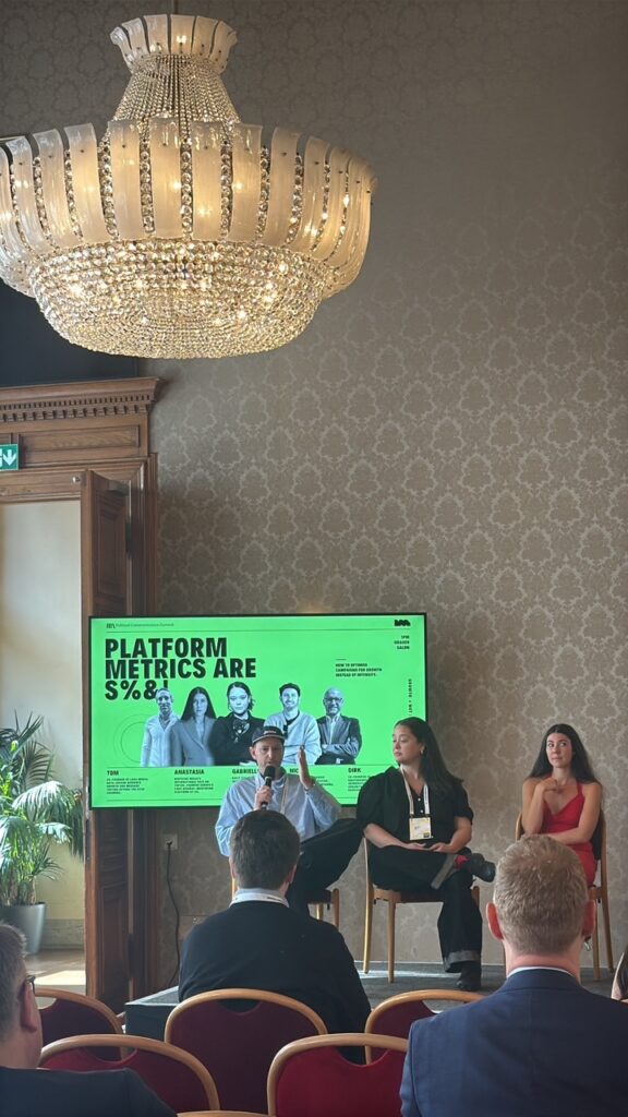 A panel discussion under a crystal chandelier, with a bright green slide behind the speakers reading “Platform metrics are S%&!”