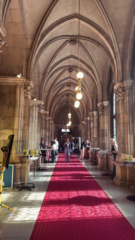 A long vaulted corridor in Vienna’s Rathaus with a red carpet, where summit attendees gather at standing tables.